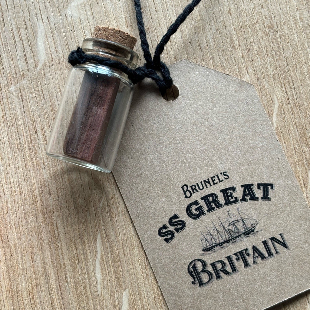 A small glass bottle with a cork stopper containing small pieces of red-brown wood from the deck of the SS Great Britain. A tag is attached to the jar which declared the origin of the wood.