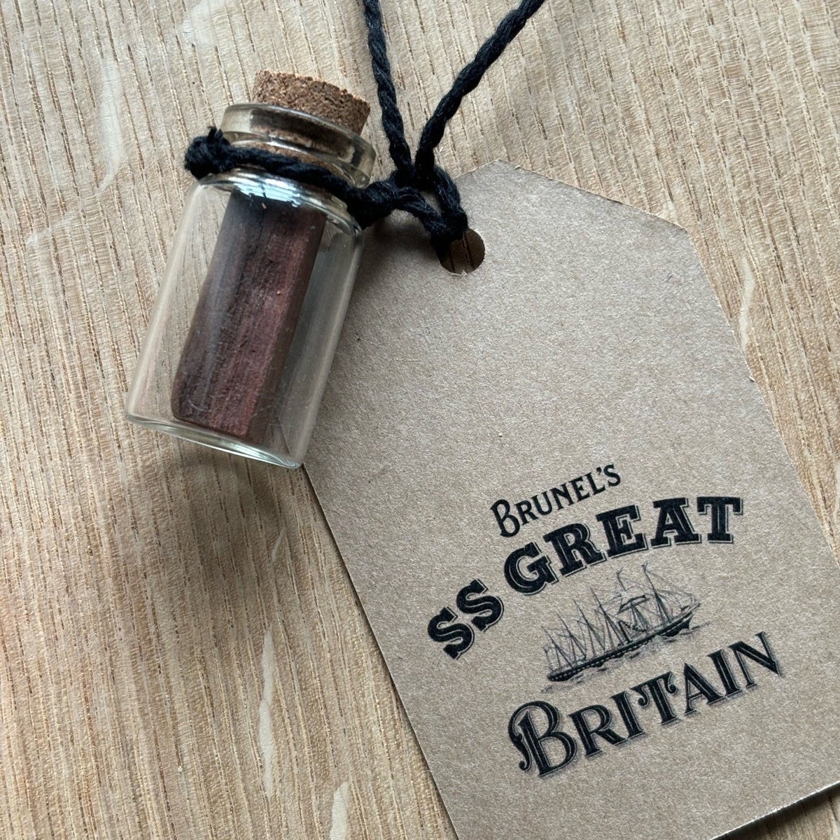 A small glass bottle with a cork stopper containing small pieces of red-brown wood from the deck of the SS Great Britain. A tag is attached to the jar which declared the origin of the wood.