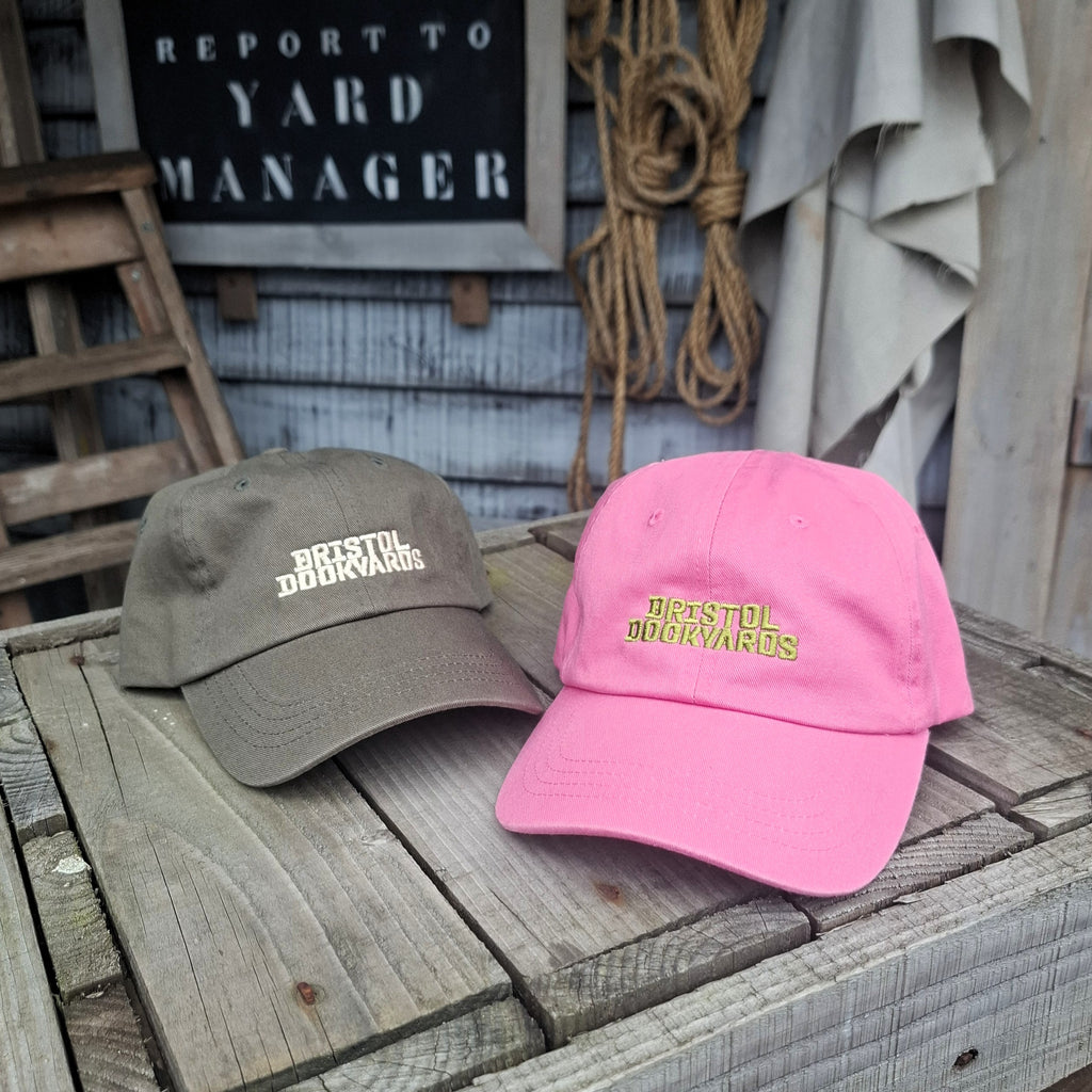 Two baseball caps, one green and one pink, with embroidered lettering reading 'Bristol Dockyards'.