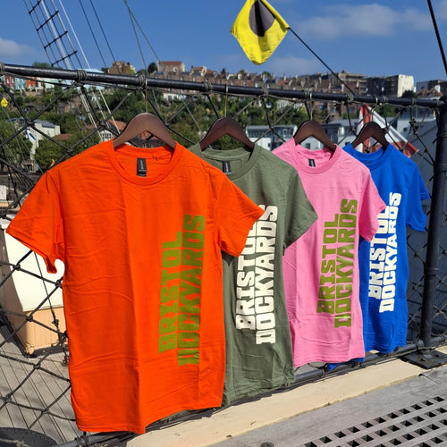 Four short-sleeve graphic T-shirts in orange, olive green, pink, and blue hang on wooden hangers outdoors on the deck of a ship. Each shirt has large vertical text reading 'BRISTOL DOCKYARDS' in contrasting colours.