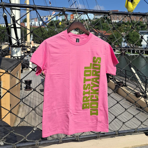 A bright pink short-sleeve t-shirt hanging on a wooden hanger over a ship's netting, featuring the words 'BRISTOL DOCKYARDS' printed vertically in green on the front.