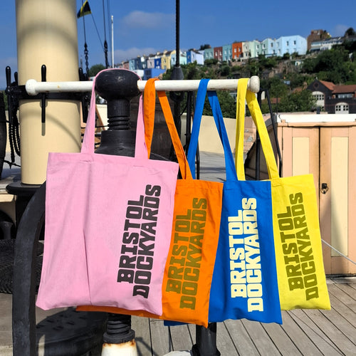 Four cotton tote bags in pink, orange, blue, and yellow hang from a white railing on a ship's deck. Each bag has the words 'BRISTOL DOCKYARDS' printed vertically in bold text.
