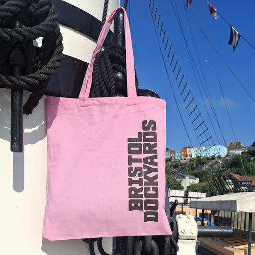 A light pink cotton tote bag with dark vertical text reading 'BRISTOL DOCKYARDS' hanging on a white ship's mast with ropes and a blue sky background.