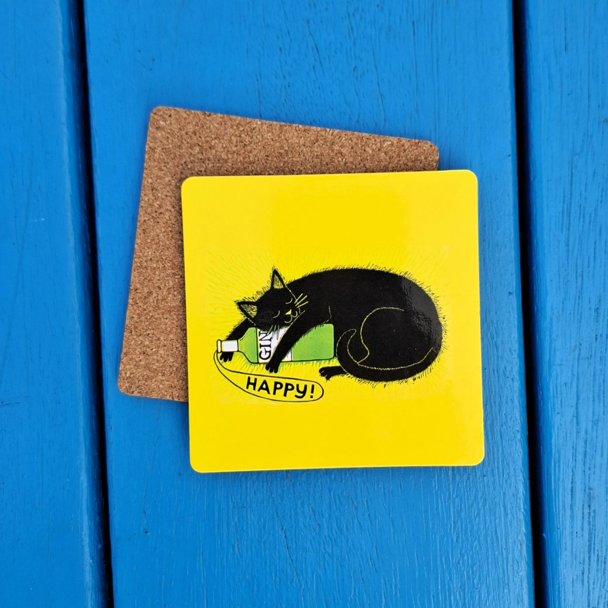 A square yellow board coaster with a black cat and gin bottle illustration. The coaster has cork backing and sits on a blue wooden surface.