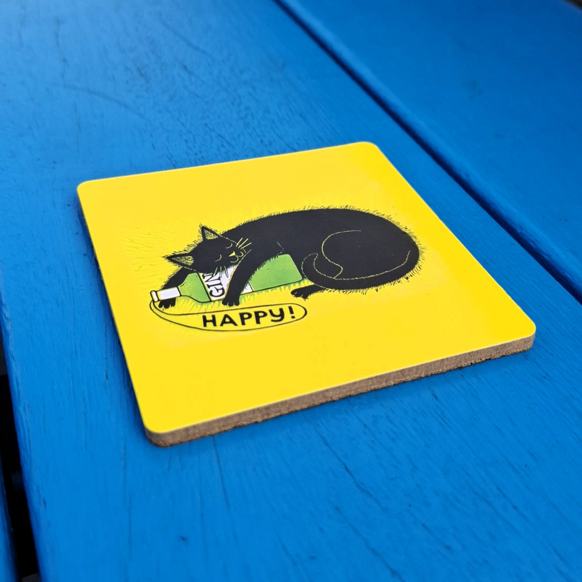 A square yellow board coaster with a black cat and gin bottle illustration. The coaster has cork backing and sits on a blue wooden surface.