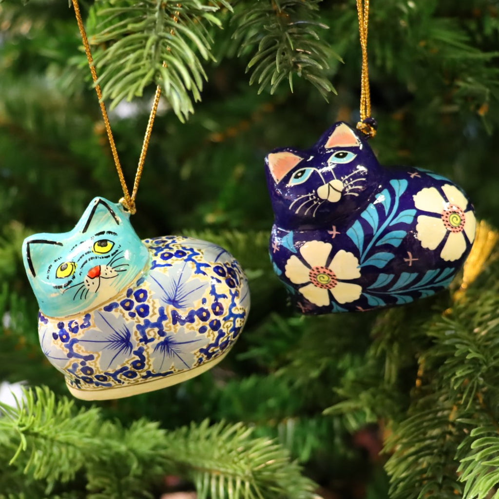 A papier mache cat decoration with a glossy finish, featuring a blue and white color scheme with floral patterns, hanging from a Christmas tree.