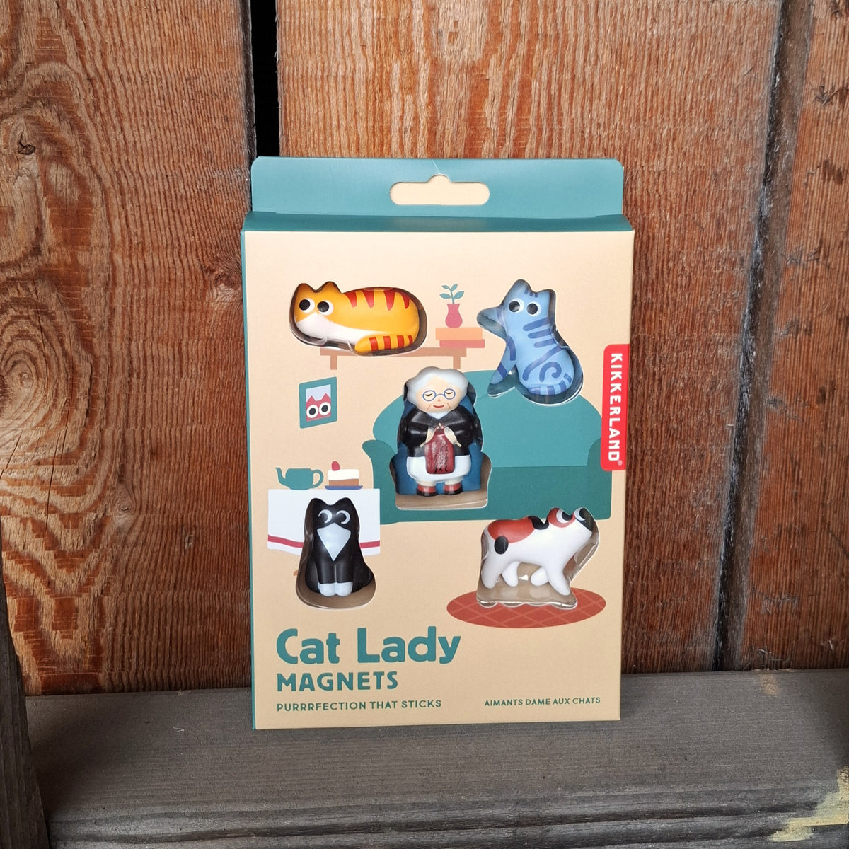 A boxed set of cat-themed magnets labeled 'Cat Lady Magnets' standing upright against a rustic wooden plank background. The packaging is cream with green accents and shows the five magnets arranged in the box.
