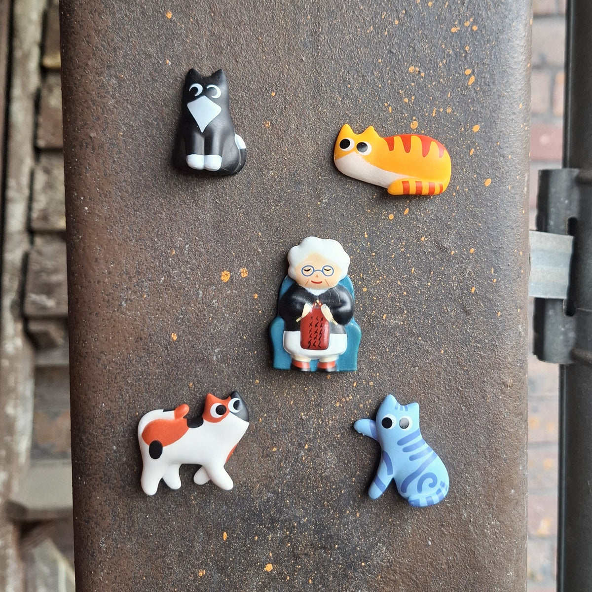 Five small magnets arranged on a dark metal surface. The set includes a black tuxedo cat, an orange tabby cat, an elderly woman knitting, a white cat with orange patches, and a blue tabby cat.