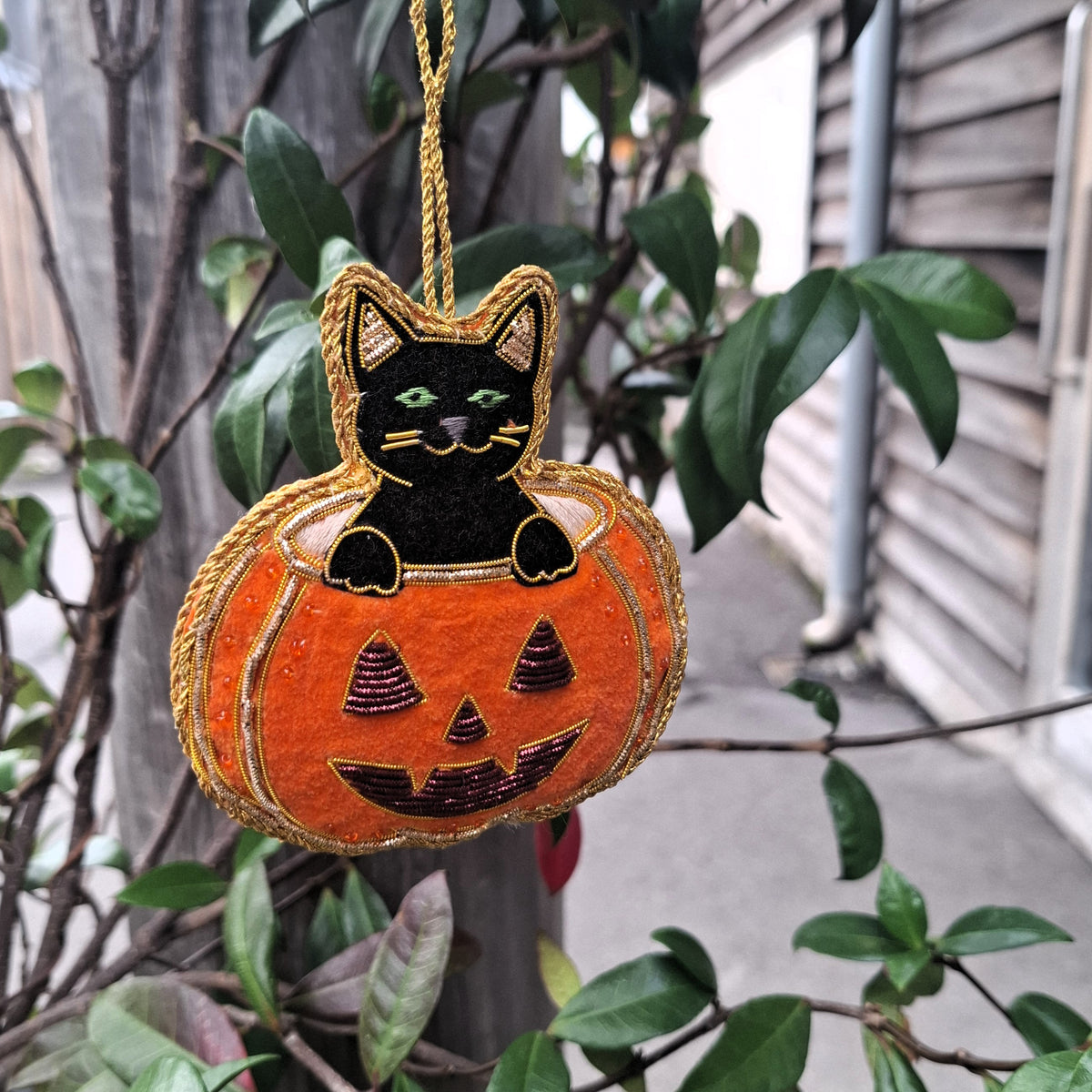 A Halloween hanging ornament shaped like a black cat sitting inside an orange jack-o'-lantern pumpkin, suspended by a gold string outdoors among green leaves and branches.