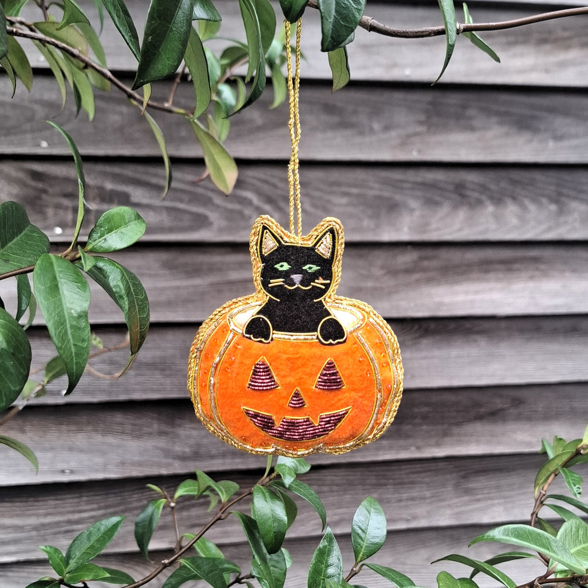 A Halloween hanging ornament shaped like a black cat sitting on an orange jack-o'-lantern pumpkin, suspended by a gold string against a gray wooden fence with green leaves in the foreground.