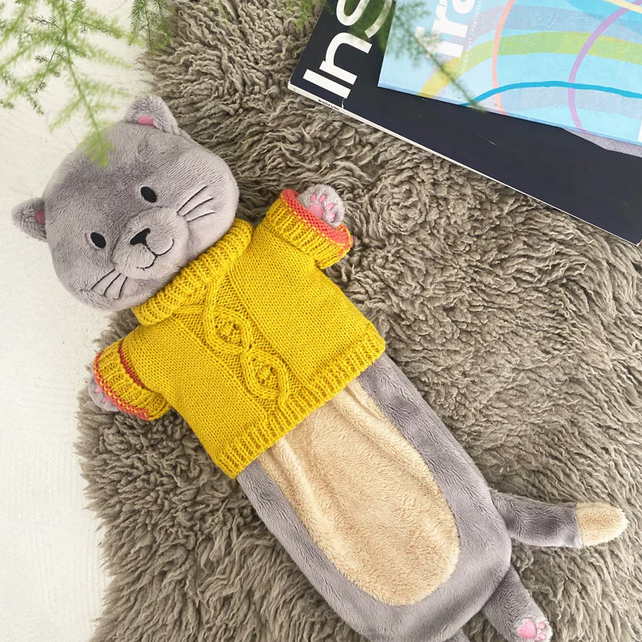 A hot water bottle cover designed to look like a grey cat with a yellow jumper, lying on a grey surface next to a book.