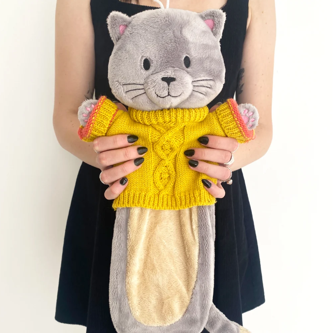 A person in a black dress holds a hot water bottle in the shape of a cat wearing a yellow knitted sweater. The cat has grey fur, pink inner ears, and a beige belly patch.
