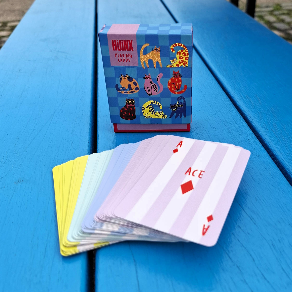 A playing cards box sits on a blue wooden surface. The box features a blue checkerboard pattern and colourful illustrations of cats. The cards are fanned out in front of the box with striped pastel designs.