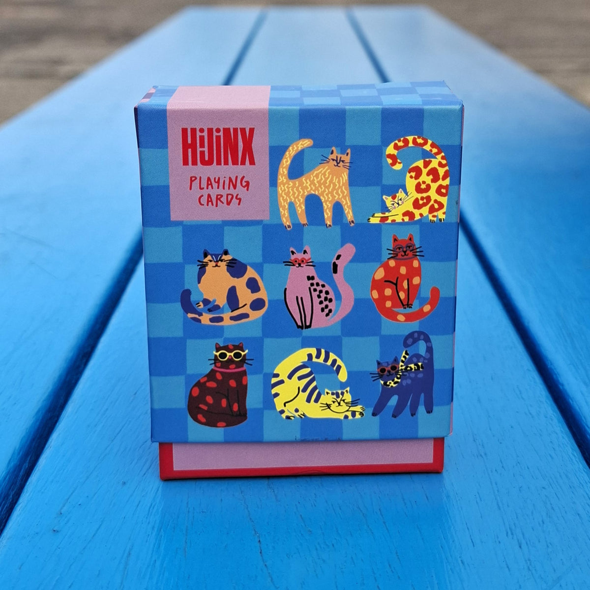 A box of playing cards sits on a blue wooden surface. The box features a blue checkerboard pattern and colourful illustrations of cats.