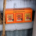 An orange Spice Kitchen Curry Collection box displayed on a rustic white wooden surface next to a thick jute rope. The box contains three tins of different spice blends: Tandoori Masala, Garam Masala, and Sri Lankan mix.