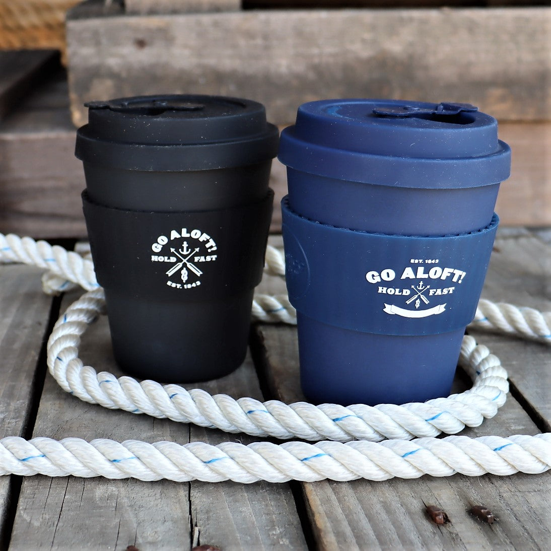 Two travel mugs, one black and one blue, with 'GO ALOFT' text on them, placed on a wooden surface with a white rope in the background.