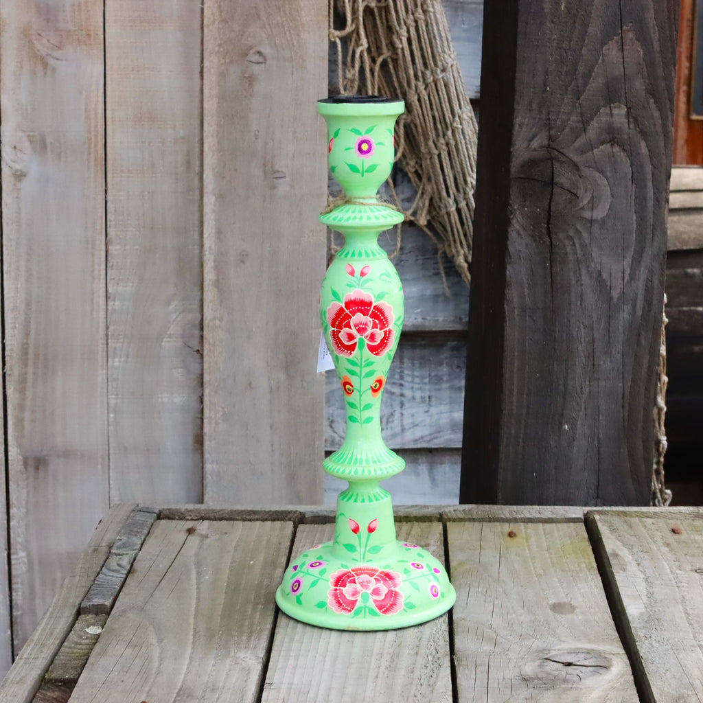 One hand-painted papier mache candlestick in mint green, decorated with red roses and green leaves, standing on a rustic wooden table against a weathered wood plank background.