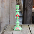 One hand-painted papier mache candlestick in mint green, decorated with red roses and green leaves, standing on a rustic wooden table against a weathered wood plank background.