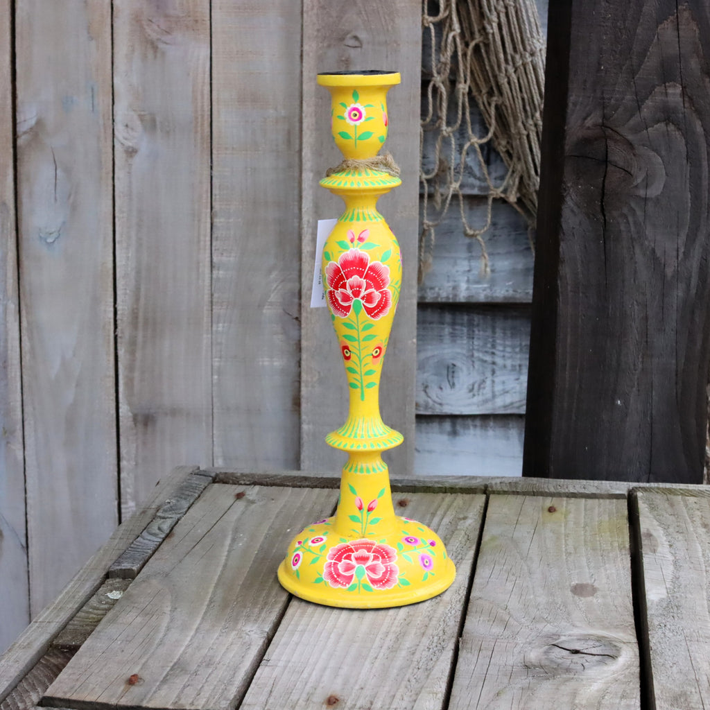 One hand-painted papier mache candlestick in yellow, decorated with red roses and green leaves, standing on a rustic wooden table against a weathered wood plank background.