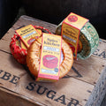 Three large tins on a wooden crate wrapped in different sari silk covers. The yellow labels read 'Spice Kitchen Indian Spice Tin, 9 Spices and Blends, Recipe book and spice spoon included'.