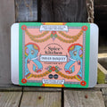 A rectangular metal tin labelled 'Spice Kitchen Indian Banquet Feasting Kit' sits on weathered wooden boards. The front of the tin features a colorful illustration of two blue elephants flanking a central peach panel with ornate gold and red decorative garlands.