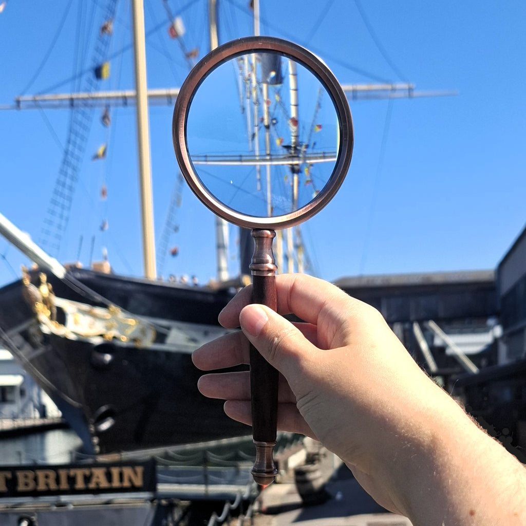 A hand holding a vintage-style magnifying glass with a copper rim and dark wooden handle. The glass is magnifying the masts of the SS Great Britain under a clear blue sky.