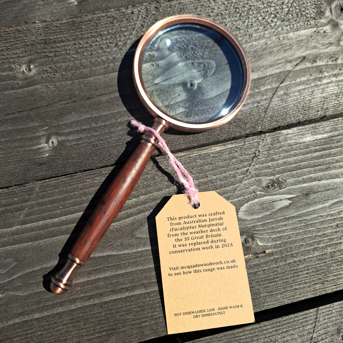 A vintage-style magnifying glass with a polished copper-toned metal rim and a dark reddish brown wooden handle, resting on a weathered wooden surface. A kraft paper tag is attached to the handle with pink string, containing information about the origin of the wood.