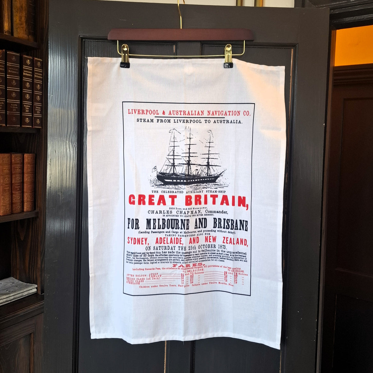A white cotton tea towel with a print of a Victorian advertising poster for the SS Great Britain's voyages to Australia in 1873. The poster design includes a line drawing of the ship and text in red and black advertising prices and destinations.