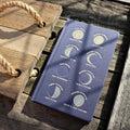 A navy clothbound journal with gold embossing featuring illustrations of the moon in different phases, placed on a wooden surface with a rope in the background.