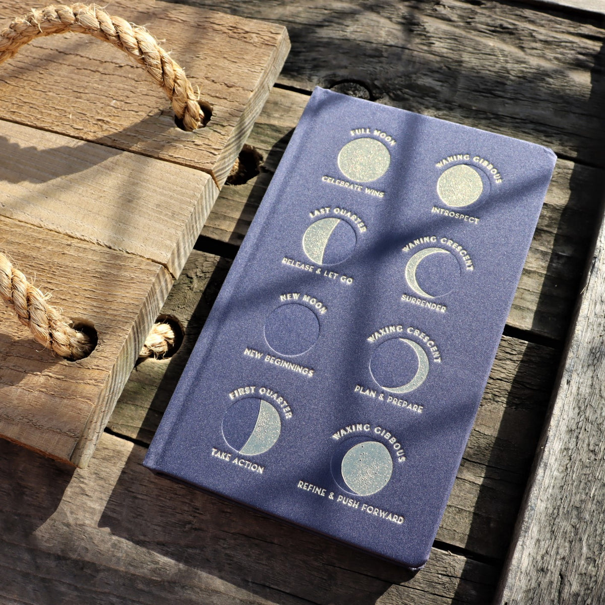 A navy clothbound journal with gold embossing featuring illustrations of the moon in different phases, placed on a wooden surface with a rope in the background.