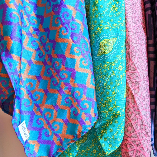 Three colourful patterned silk fabrics hanging side-by-side. The leftmost fabric features a turquoise, purple, and orange geometric pattern.