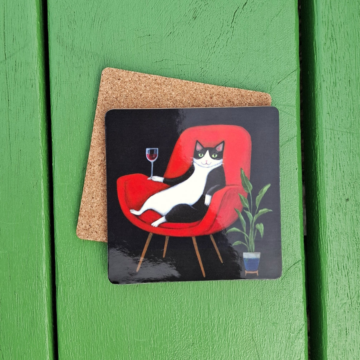 A shiny black board coaster featuring an illustration of a black-and-white cat lounging in a red armchair and holding a glass of red wine. The coaster is backed with cork and sitting on a green wooden background.