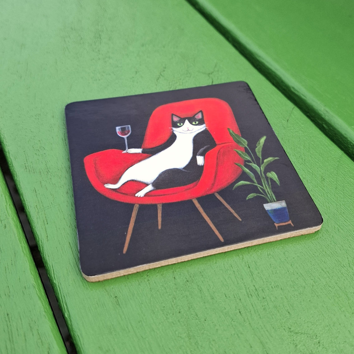 A shiny black board coaster featuring an illustration of a black-and-white cat lounging in a red armchair and holding a glass of red wine. The coaster is backed with cork and sitting on a green wooden background.