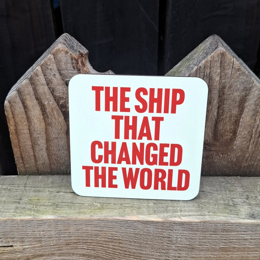 A pale green coaster with 'The Ship That Changed The World' orange text on a wooden surface