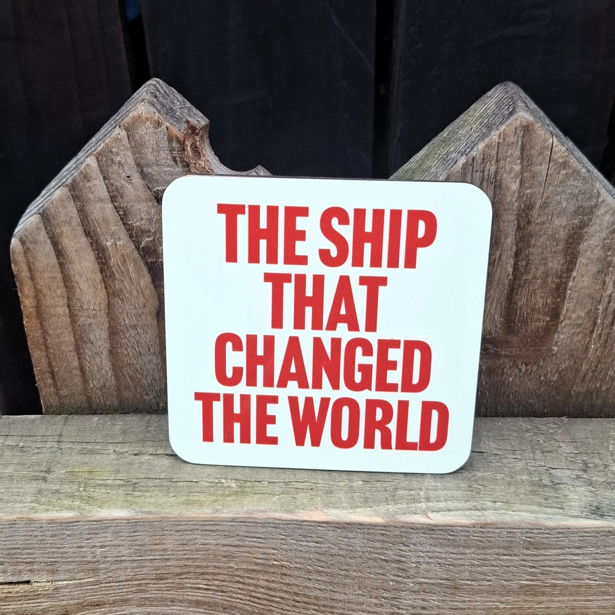 A pale green coaster with 'The Ship That Changed The World' orange text on a wooden surface