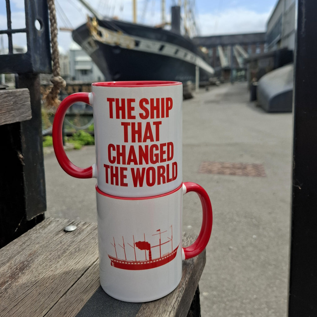 Two mugs are stacked on top of each other, showing the two sides of the design. One side features the text 'the ship that changed the world' in red lettering. The other side has an illustration of the SS Great Britain in red. The mugs are stacked in the dockyard with the real SS Great Britain in the background.