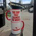 Two mugs are stacked on top of each other, showing the two sides of the design. One side features the text 'the ship that changed the world' in red lettering. The other side has an illustration of the SS Great Britain in red. The mugs are stacked in the dockyard with the real SS Great Britain in the background.
