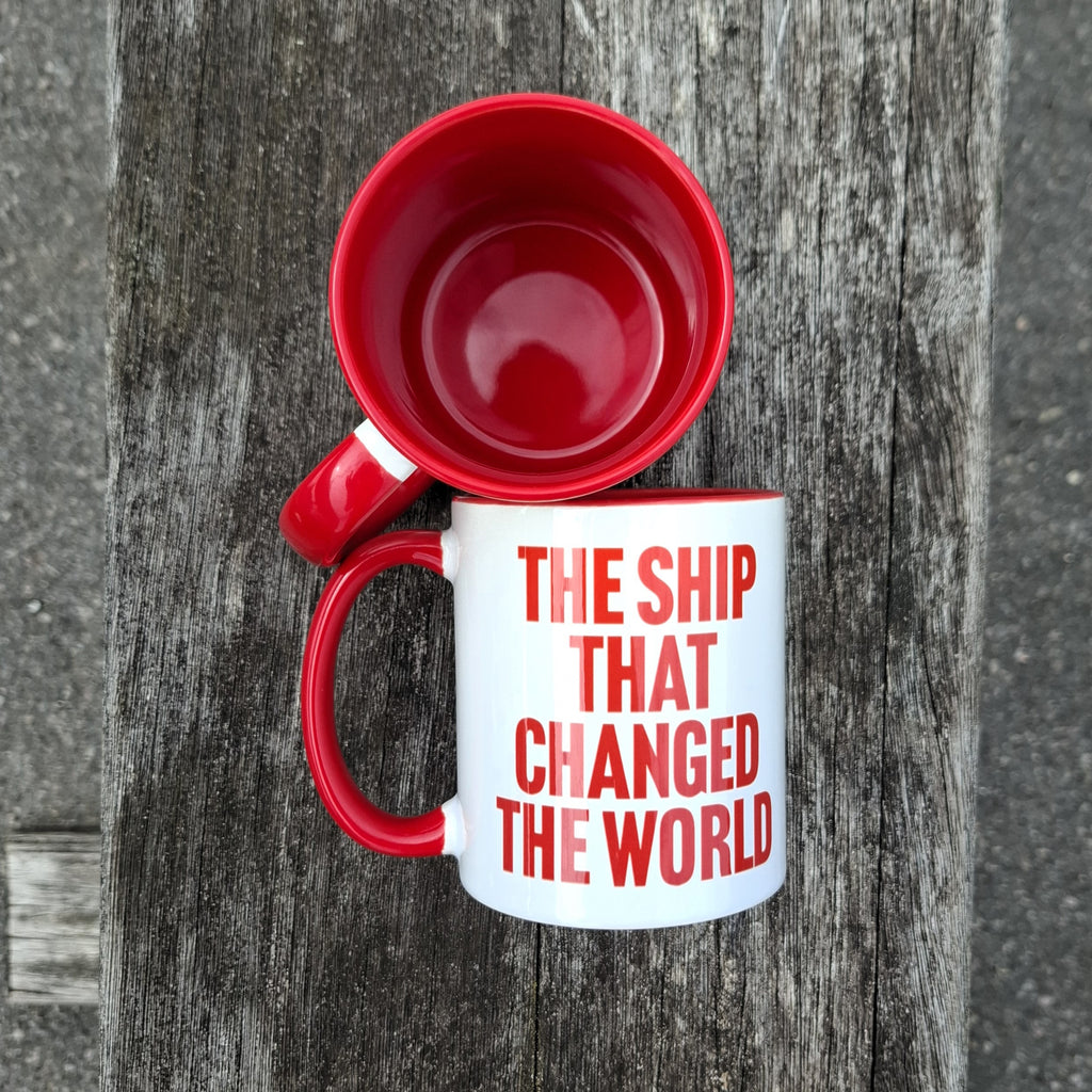 A view from above of two mugs, one standing up, one lying on its side. The mug standing up shows the red interior of the mug, while the one lying down shows the text 'The Ship That Changed the World'.