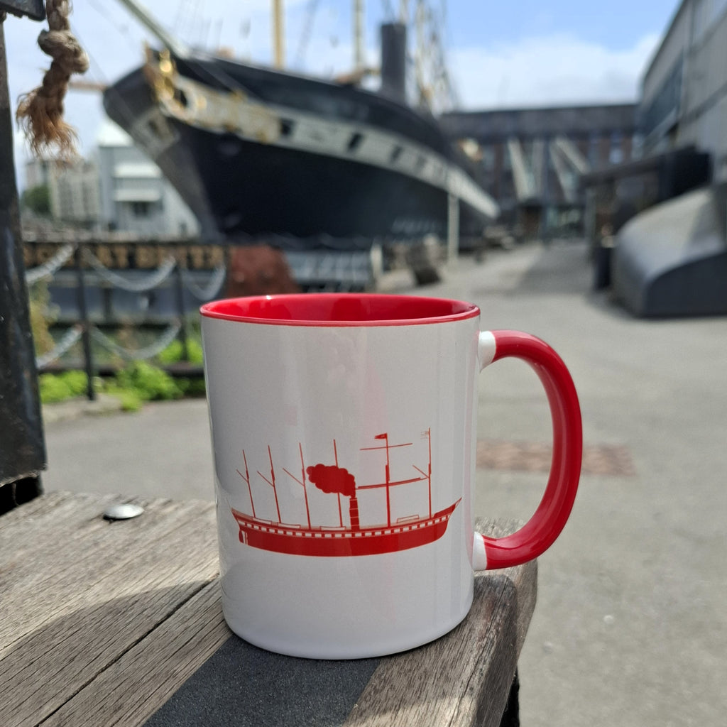 A white ceramic mug with a red handle and red interior, featuring a line drawing of the SS Great Britain in red. The mug sits on a wooden surface with the SS Great Britain in the background.
