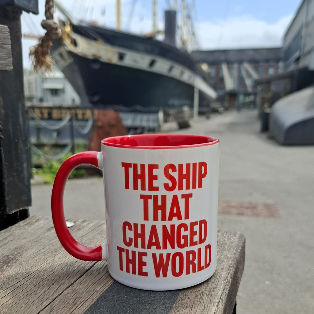 A white ceramic mug with a red handle and red interior, printed with the phrase 'THE SHIP THAT CHANGED THE WORLD' in bold red letters, sitting on a wooden surface with the SS Great Britain in the background.