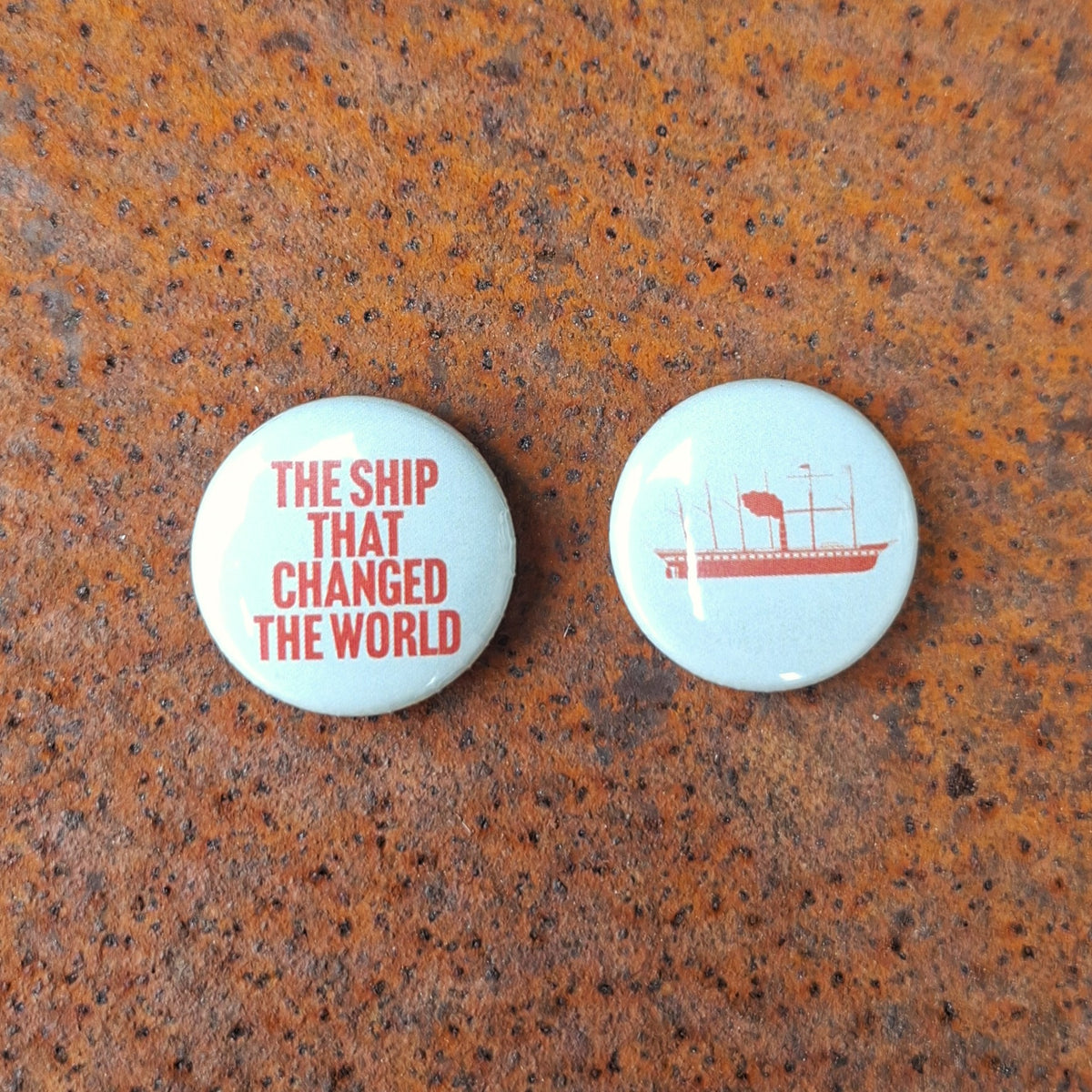 Two round pale green button badges with red printed graphics, placed on a rusty metal surface. One badge reads 'THE SHIP THAT CHANGED THE WORLD' and the other shows a red ship illustration.