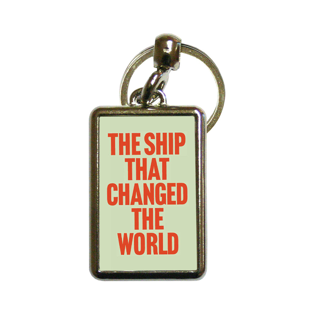 A rectangular metal keychain with a light green background and bold red text reading 'THE SHIP THAT CHANGED THE WORLD' on a white background.