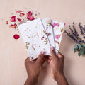 A person's hands holding handmade paper with pressed flowers, alongside scattered dried flowers.